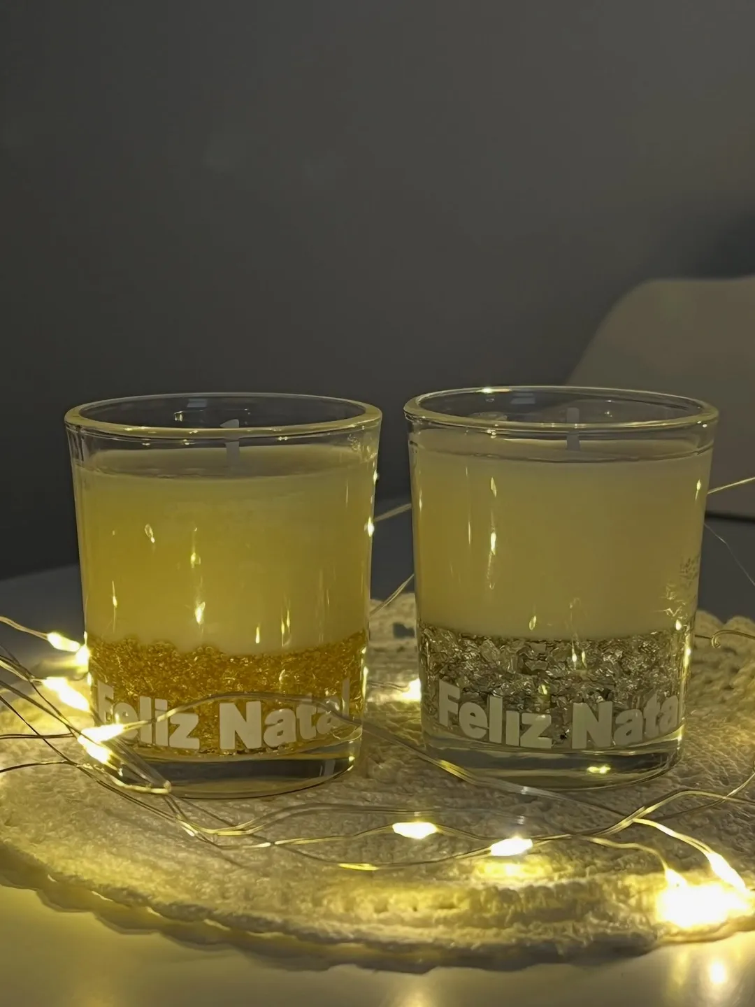 Dois copos de vela IlumiAromas com a gravação Feliz Natal, cercados de luzes